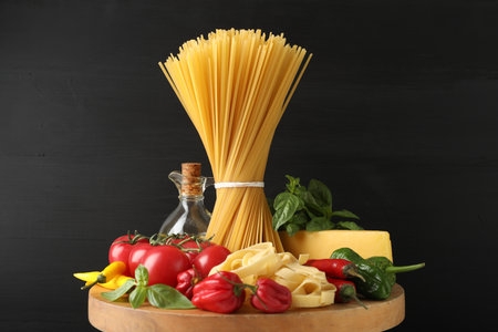 Different types of raw pasta and products on wooden board against black background, closeupの写真素材