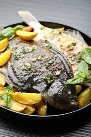 Tasty roasted flounder fish with herbs and potato on black table, closeup. Homemade seafood dish