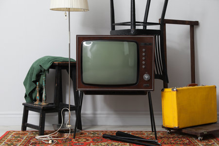 Retro TV on stand, black chair, suitcase and lamp near white wall. Used stuffの写真素材