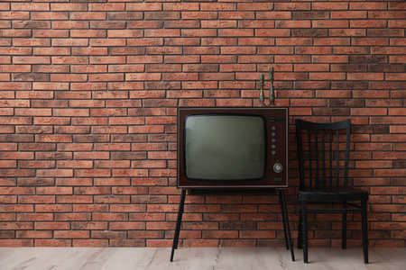 Retro TV on stand and black chair near red brick wall, space for text. Used stuffの写真素材