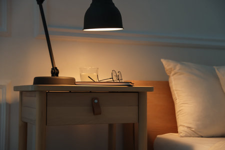 Bedside table with lamp, notebook, glasses and glass of water near bed indoors