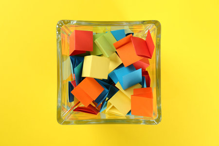 Colorful paper pieces in glass jar on yellow background, top view