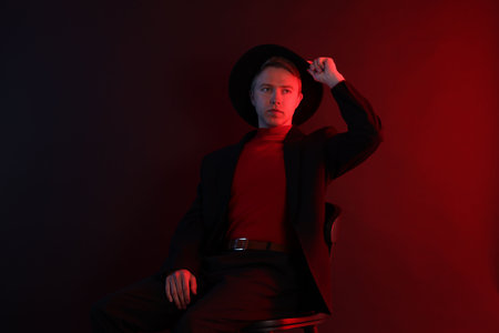 Handsome man wearing suit and hat sitting on chair in neon lights against dark backgroundの写真素材