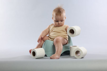 Cute little baby sitting on potty and rolls of toilet paper against light grey background