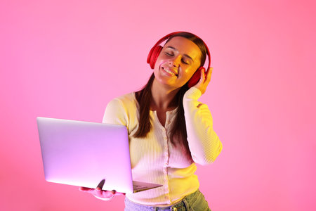 Smiling woman in headphones with laptop in neon lights against pink backgroundの写真素材