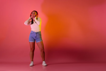 Smiling woman in headphones listening to music in neon lights against pink background. Space for textの写真素材