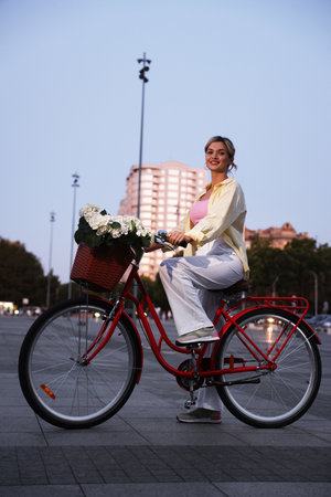 Beautiful woman riding bicycle with basket of flowers on city streetの写真素材