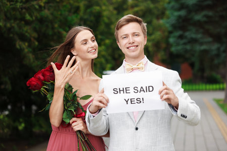 Smiling man holding sheet of paper with text She Said Yes and his happy beloved with bouquet of roses showing ring on engagement day outdoorsの写真素材