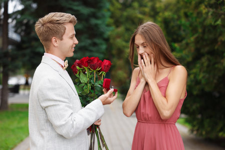 Smiling man with engagement ring and bouquet of roses making marriage proposal to his surprised...