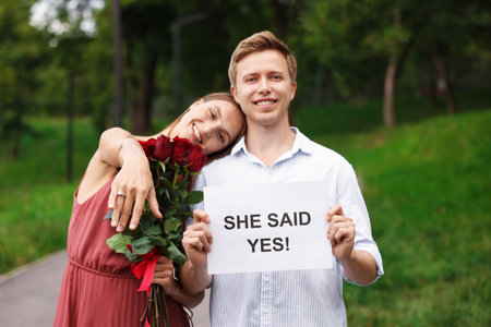 Smiling man holding sheet of paper with text She Said Yes and his excited beloved with bouquet of roses showing ring on engagement day outdoorsの写真素材