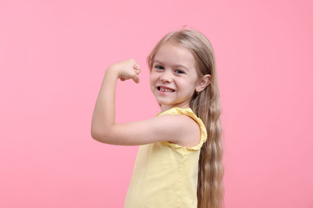 Strength. Happy little girl showing her biceps on pink background