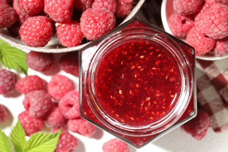 Delicious raspberry jam in glass jar, fresh berries and mint on white table, flat lay