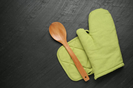 Green oven glove, wooden spoon and potholder on dark textured table, flat lay. Space for textの写真素材