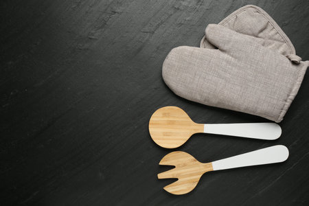 Gray oven glove and other kitchen utensils on dark textured table, flat lay. Space for text