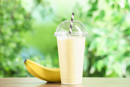 Tasty milkshake in plastic cup and banana on wooden table outdoorsの写真素材