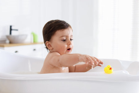 Cute little baby with rubber duck bathing in tub at home