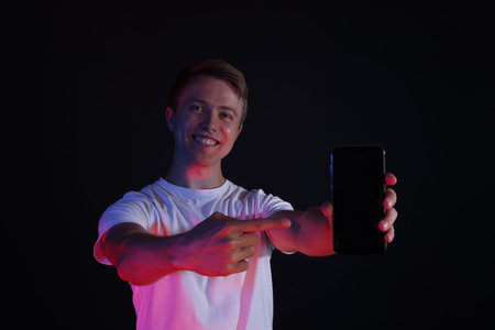 Smiling man pointing at smartphone in neon lights against black background