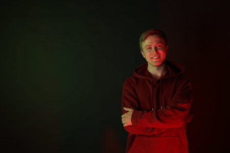 Portrait of smiling man wearing hoodie with crossed arms in neon lights against black background....