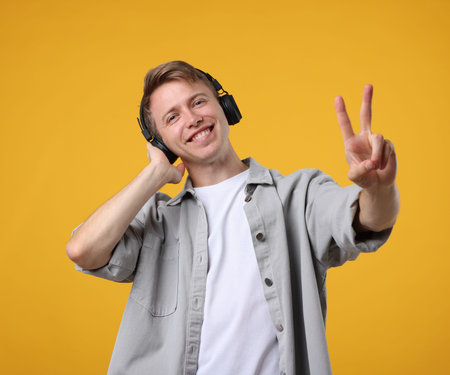 Happy man listening to music in headphones and showing peace sign on yellow backgroundの写真素材