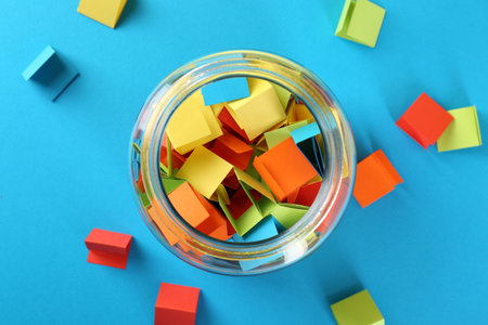 Colorful paper pieces in glass jar on light blue background, top view