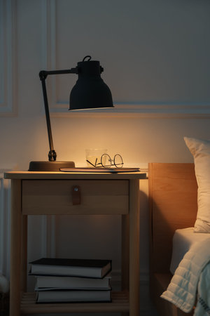 Bedside table with lamp, notebook, glasses and glass of water near bed indoors