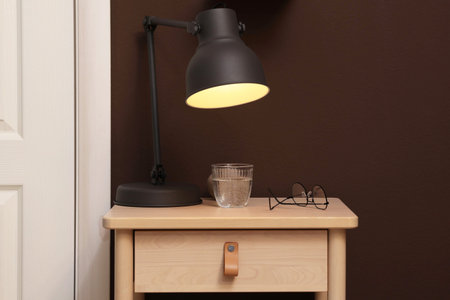Wooden side table with glass of water, lamp and glasses near brown wall indoors