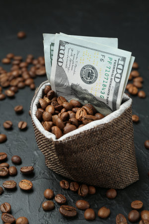 Dollar banknotes and roasted coffee beans in sack on dark textured table, closeup