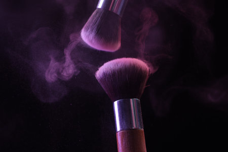 Makeup brushes with purple eye shadow in neon lights on black background, closeup
