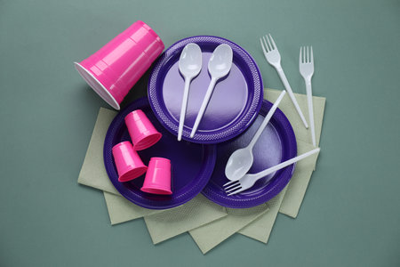 Set of disposable plastic tableware and paper napkins on green background, flat lay
