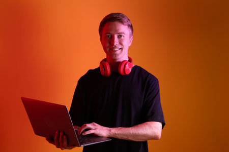 Portrait of smiling man with headphones using laptop in neon lights against orange backgroundの写真素材