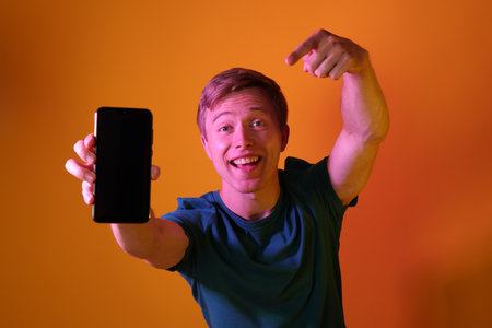 Happy man pointing at smartphone in neon lights against orange backgroundの写真素材