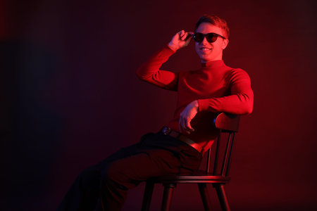 Smiling man wearing sunglasses sitting on chair in neon lights against dark backgroundの写真素材