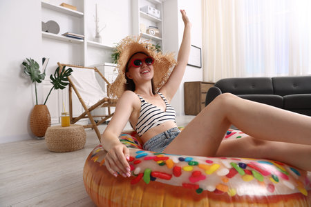 Happy woman in sunglasses and straw hat with inflatable ring having fun on floor at home. Dreaming about vacation at seaの写真素材