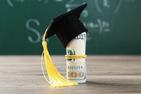 Scholarship. Rolled dollar banknotes with graduate hat on wooden table against green chalkboard, closeupの写真素材