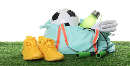 Stylish gym bag with sports equipment, towel, bottle and sneakers on green grass against white backgroundの写真素材
