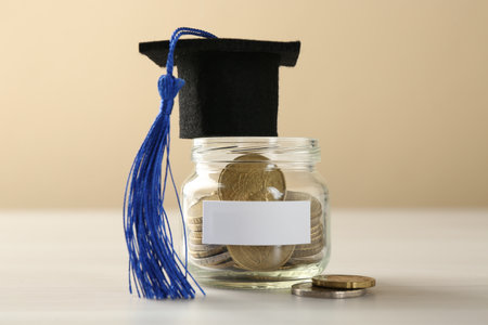 Scholarship. Jar with graduate hat and coins on white wooden table against beige background, closeupの写真素材