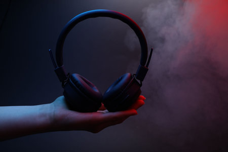 Woman holding wireless headphones on dark background with color lights and smoke, closeupの写真素材