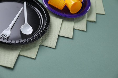 Set of disposable plastic tableware and paper napkins on green background, closeup. Space for text
