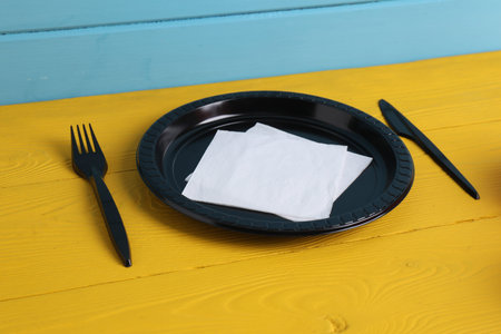 Set of disposable plastic tableware on yellow wooden table against light blue background, closeup