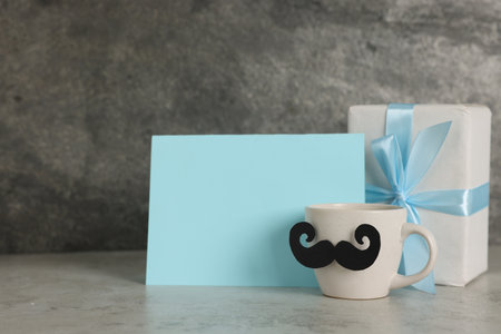 Happy Father's Day. Cup with paper moustache, gift box and card on light gray table, space for textの写真素材