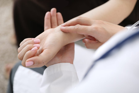 Doctor checking woman's pulse with fingers indoors, closeup