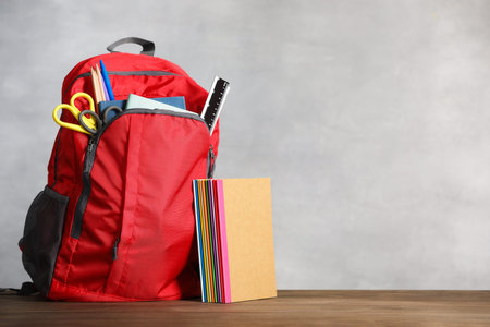 Red backpack with different school stationary on wooden table against gray background. Space for...