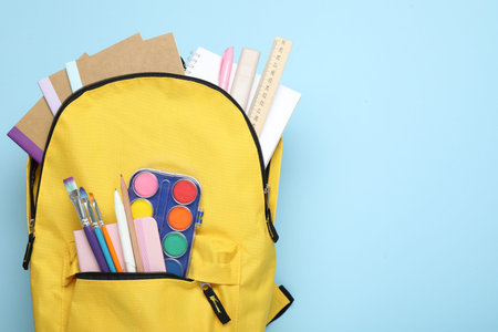 Yellow backpack with different stationery on light blue background, top view. Space for text