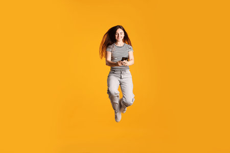 Happy woman with beautiful long hair using smartphone and jumping on orange backgroundの写真素材