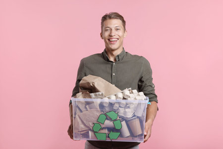 Recycling. Smiling man holding plastic box with different waste paper on pink background