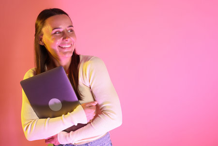 Smiling woman with laptop in neon lights against pink background. Space for textの写真素材