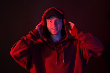 Portrait of smiling man wearing hoodie, baseball cap and headphones in neon lights against black backgroundの写真素材