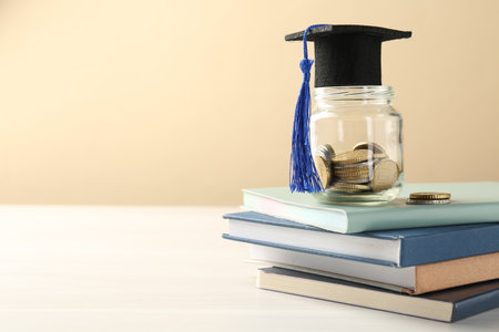 Scholarship. Jar with graduate hat, coins and books on white wooden table against beige background, space for textの写真素材