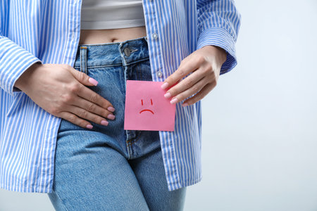 Woman holding sticky note with drawn sad face and suffering from cystitis symptoms on light grey background, closeup. Space for textの写真素材