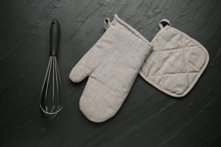 Grey oven glove, potholder and whisk on dark textured table, flat layの写真素材